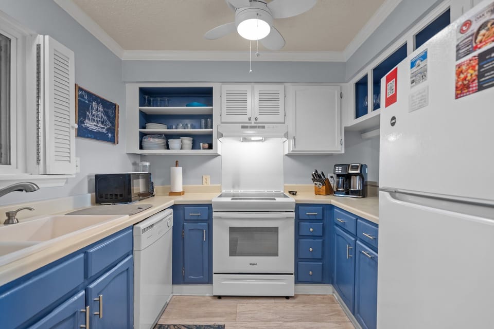 Oak Island, NC kitchen with blue lower cabinets, white appliances, stove, sink, dishwasher, microwave, coffee maker, ceiling fan, and nautical décor.