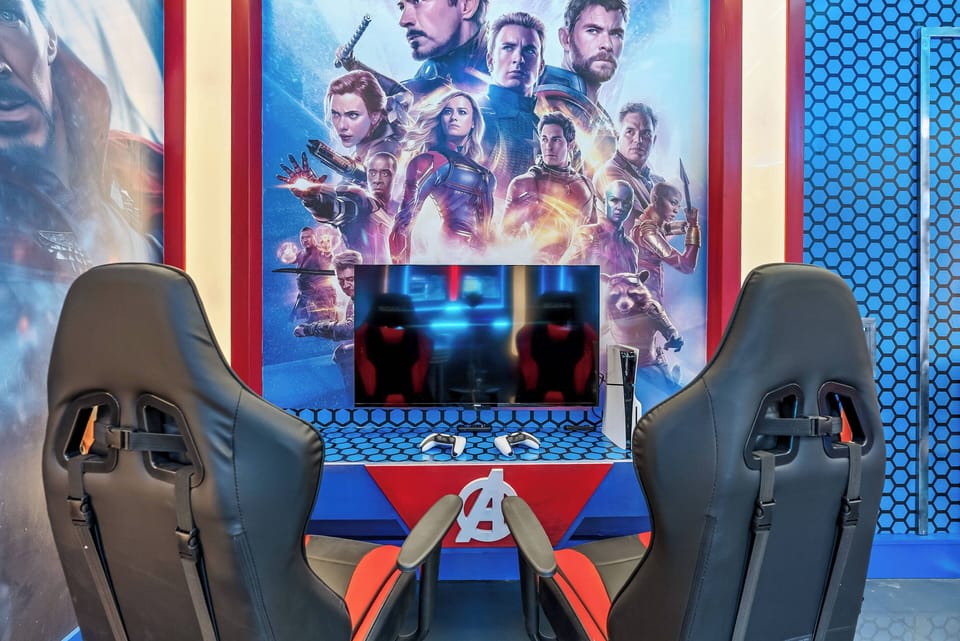Welcome to our vibrant gaming lounge, featuring sleek racing chairs and a wall of iconic superhero artwork. The bold blue and red accents create an energetic atmosphere, ideal for hours of relaxation and fun gaming.