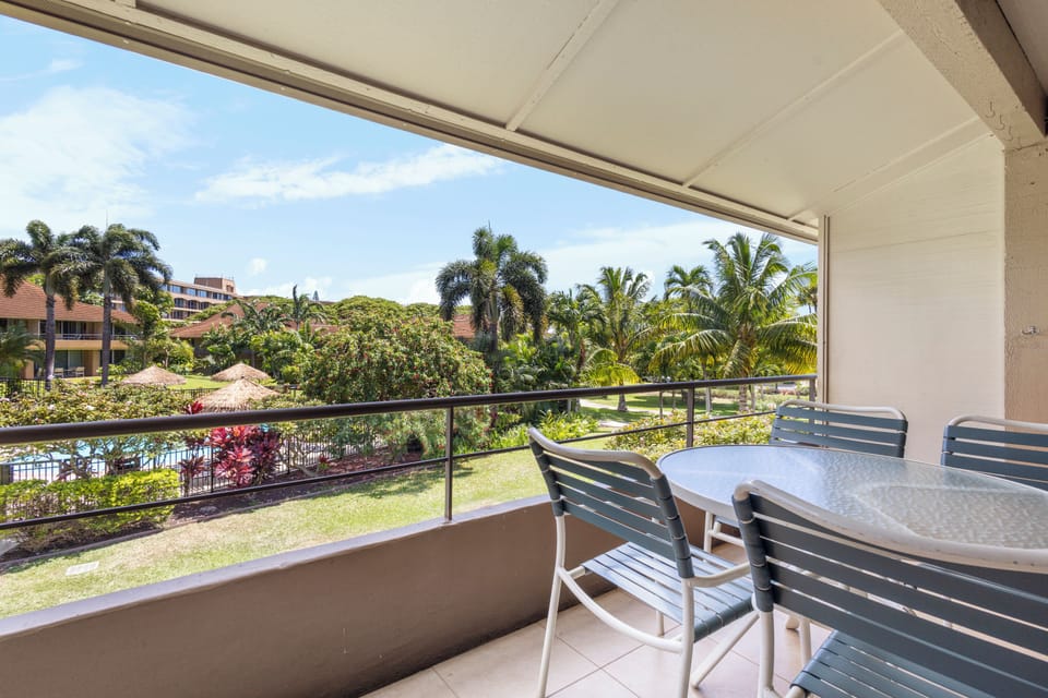 A modern balcony with patio seating overlooks lush tropical gardens and palm-filled landscaping, surrounded by the quiet property grounds and neighboring buildings.