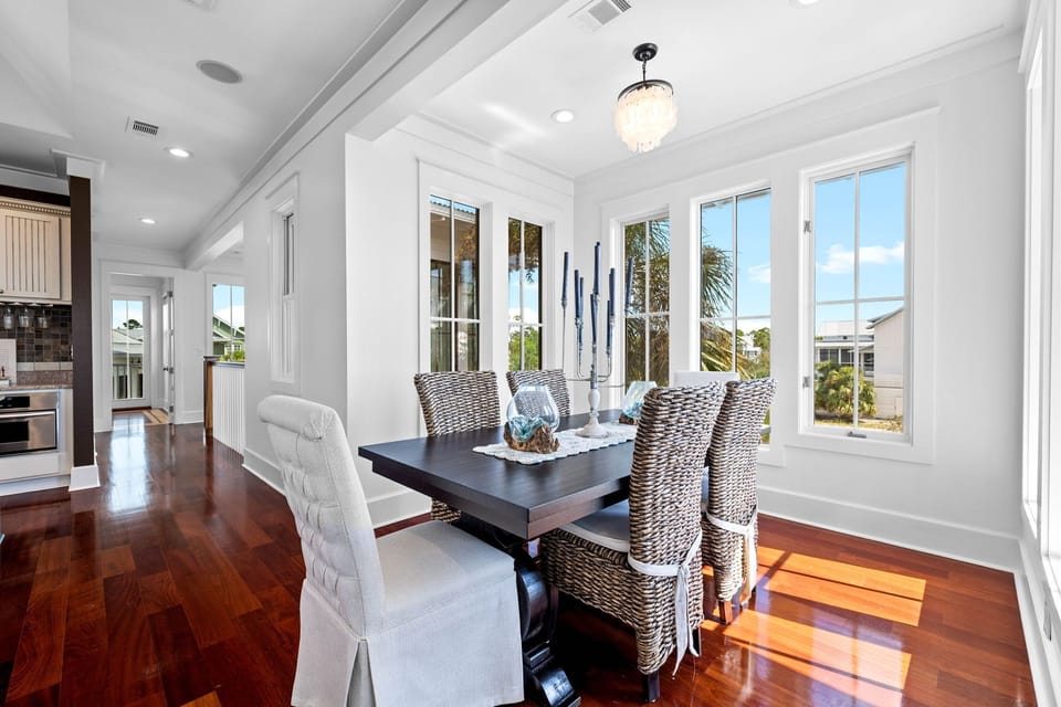 Gather around your elegant dining table where warm hardwood floors meet coastal charm and windows frame tropical views.