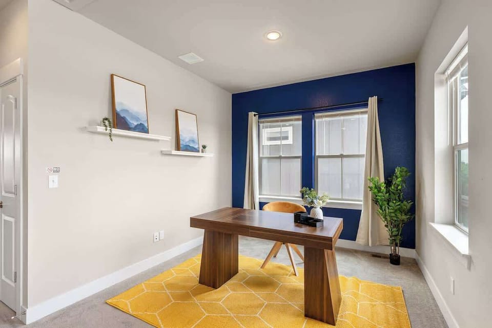 Cozy and modern home office with a wooden desk, comfortable chair, and bright yellow rug. Features a bold navy accent wall with two large windows for natural light, minimalist wall shelves with art, and a touch of greenery for a refreshing atmosphere