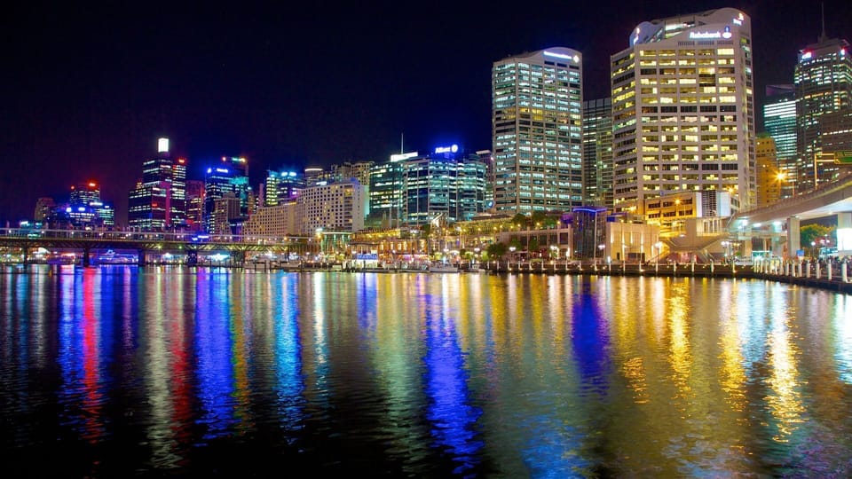 A vibrant nocturnal view of the city fronting the harbor, where illuminated skyscrapers bleed into the dark water, creating a dazzling canvas of bleeding reds, blues, and golds.