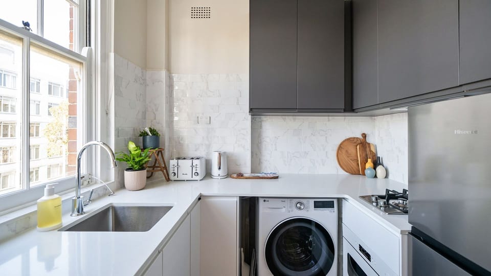 A masterclass in small-space efficiency, featuring high-contrast charcoal and white cabinetry, a built-in washing machine, a gas cooktop, and a bright window over the under-mount sink.