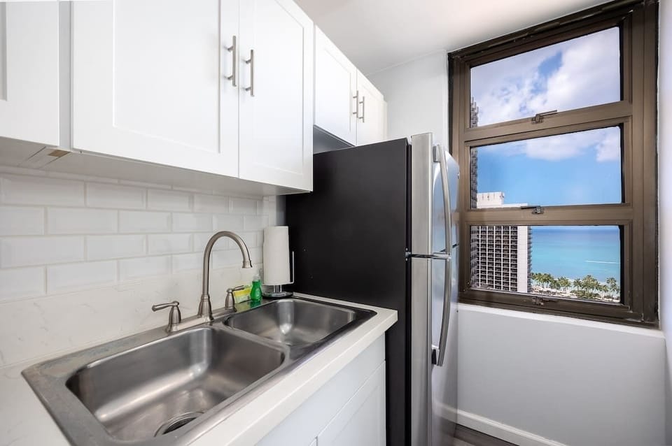 This modern kitchen offers white cabinetry, a convenient double sink, and a full-sized refrigerator, all complemented by a window providing refreshing natural light and peeks of the ocean view.