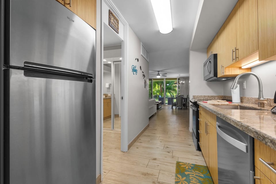 Your fully equipped kitchen features stainless steel appliances, granite countertops, and modern cabinetry. The open layout flows seamlessly into the living area, where tropical garden views await through large windows.