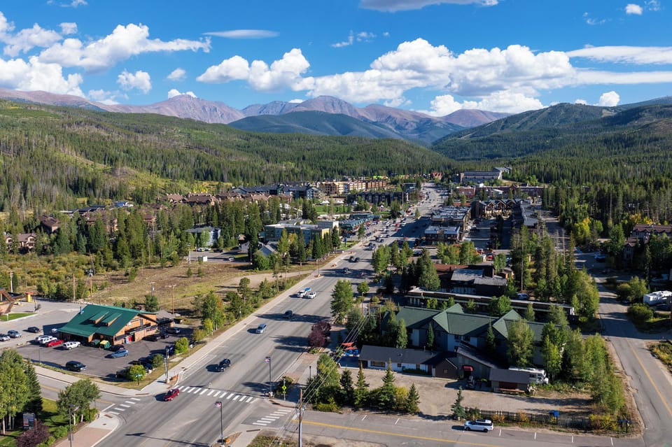 Scenic aerial shot of a charming mountain town with forests, winding roads, and snow capped peaks in the distance.