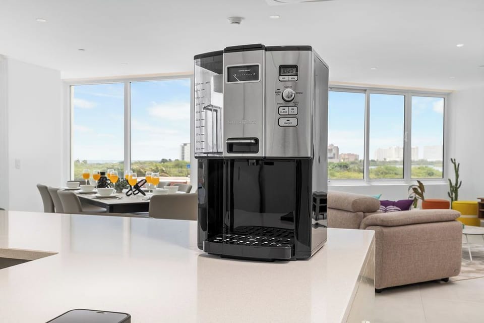 Modern living at your fingertips—a sleek coffee maker on the counter, with sun-drenched dining and lounging spaces framing peaceful city park views beyond.
