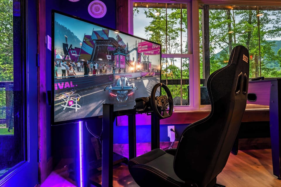 Gaming setup with racing simulator and scenic forest views through large windows.