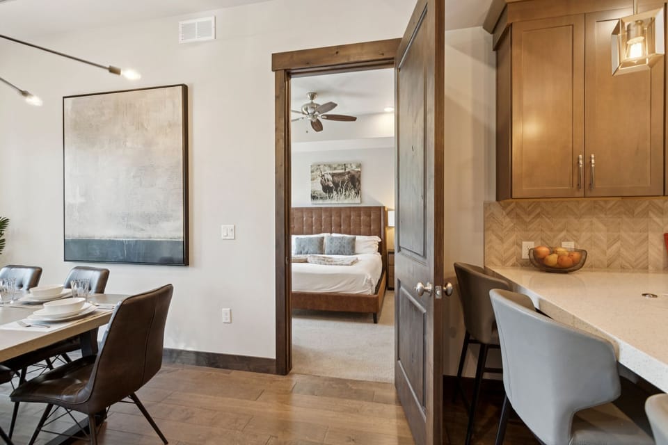 An open-concept living space connects a modern dining area, a warm kitchen with a breakfast bar, and a glimpse of a cozy bedroom.