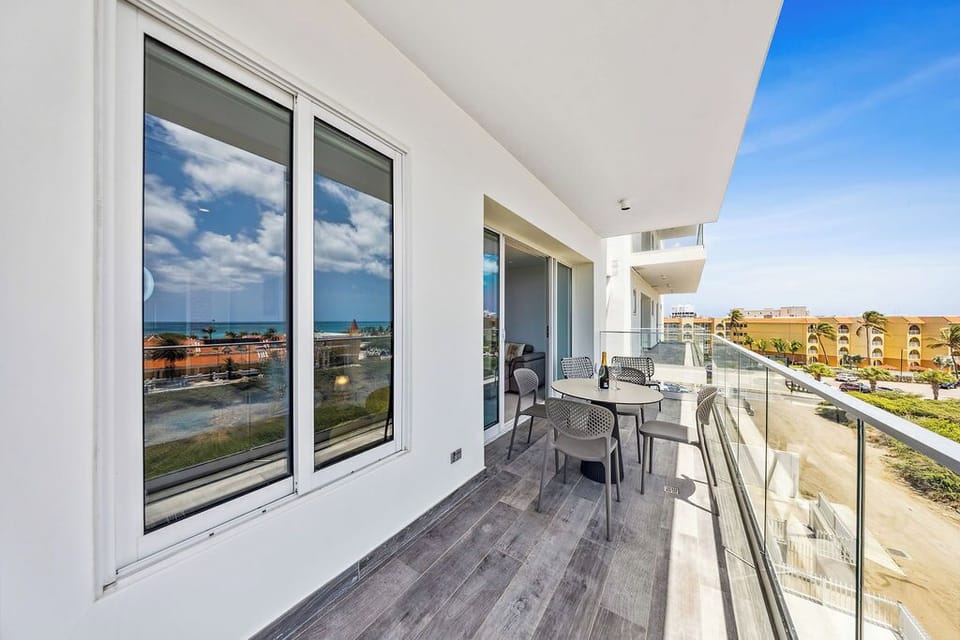 The spacious balcony offers modern seating and glass railings, framing views of the ocean, nearby buildings, and lush tropical surroundings—a perfect spot for morning coffee or sunset drinks.