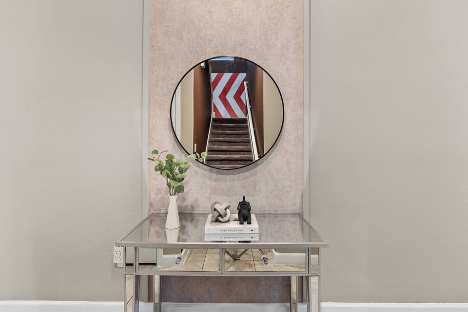 Welcome to a stylish entryway with a chic silver console table, decorative accents, and fresh flowers. A round mirror brightens the space, and captivating artwork guides you to a warm stairway, creating the perfect mood for your.