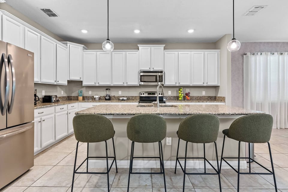 Enjoy a stylish kitchen featuring elegant white cabinetry and a spacious granite island, perfect for meals or coffee. The cozy green bar stools add warmth, while natural light from the nearby window brightens the space.