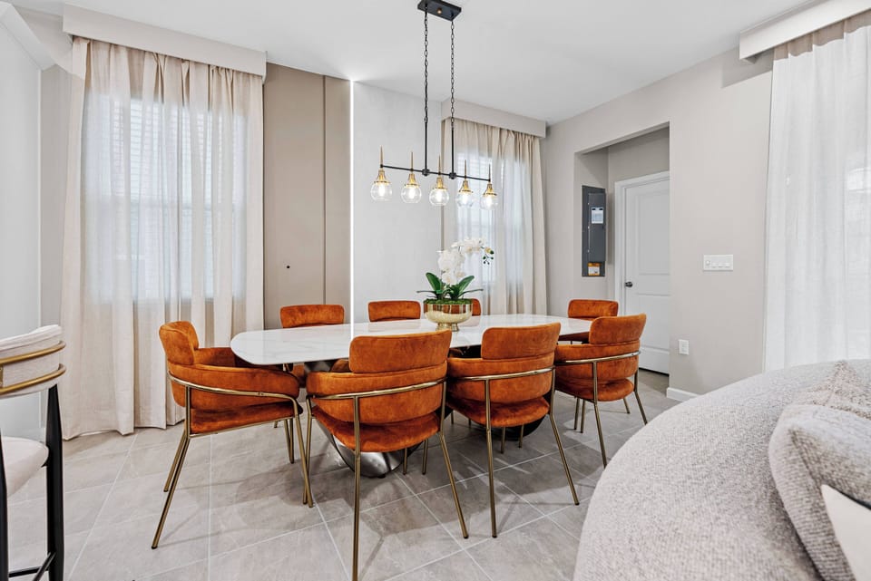 Enjoy meals and conversations at our chic round dining table, surrounded by plush rust-colored chairs that create a lively atmosphere. Floor-to-ceiling curtains frame natural light, enhancing the inviting decor.