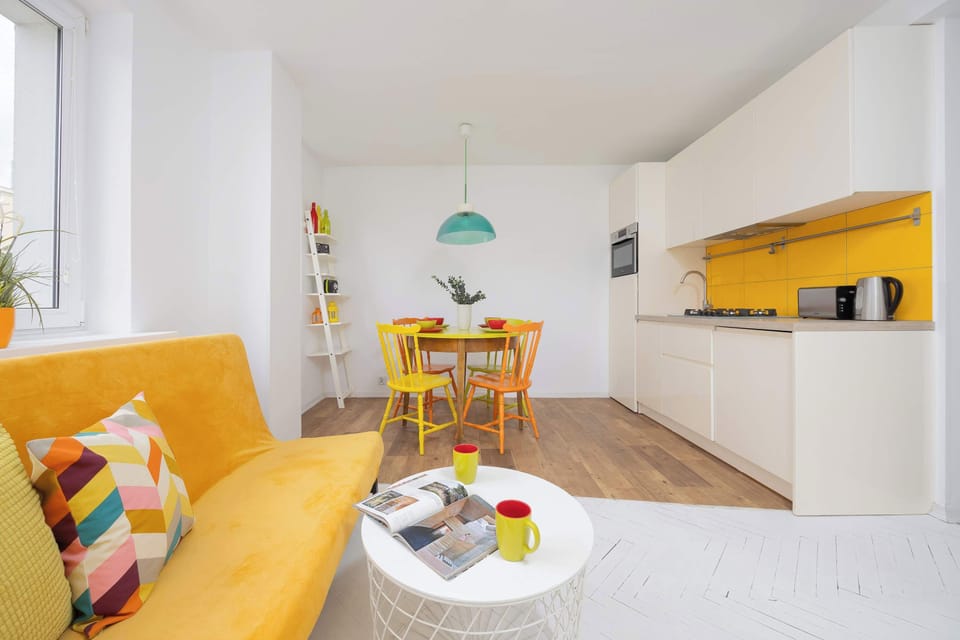 A sunny kitchen and living area with a mustard yellow sofa, colorful pillows, and white cabinetry creating a vibrant atmosphere.
