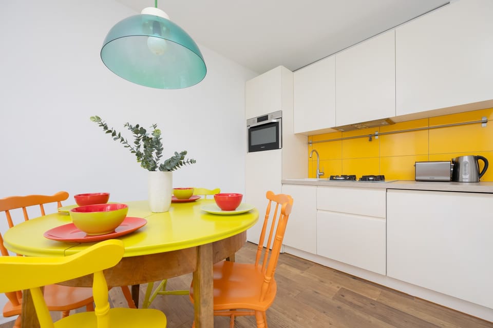 A minimalist dining corner with a yellow round table, bright green pendant light, and colorful chairs.