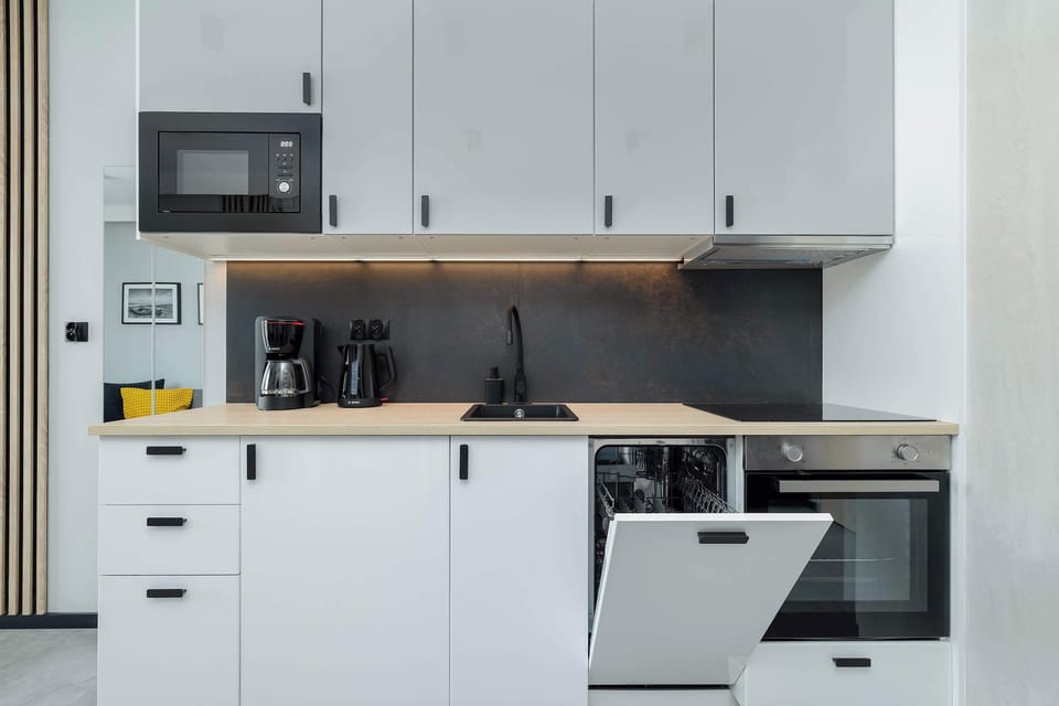 A functional kitchenette with sleek white and gray cabinets, a dark panel over the countertop and built-in appliances - such as a dishwasher, oven and microwave - offers modern amenities in a compact, aesthetically pleasing space, ideal for everyday 