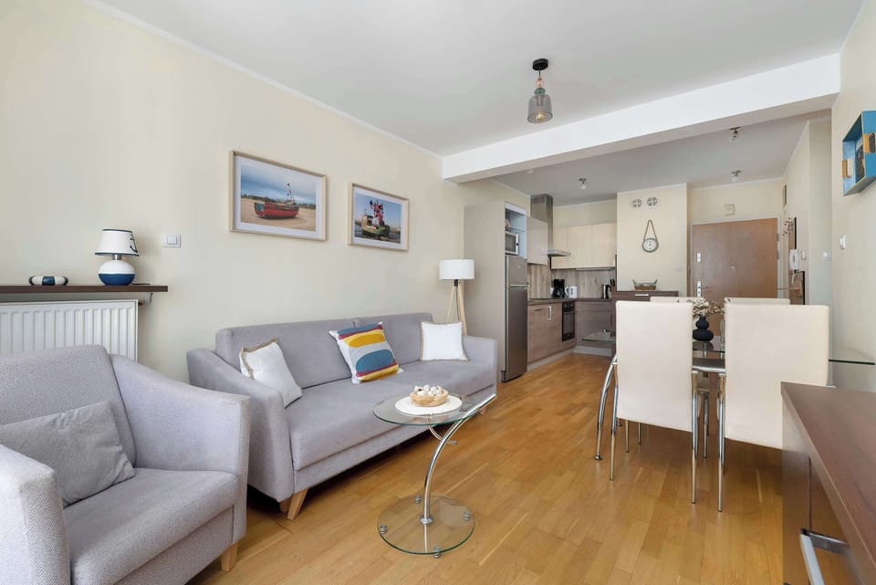 An open-plan living space combines a sofa area with a dining table and kitchen. The room has bright walls, wooden floors, and a comfortable layout.