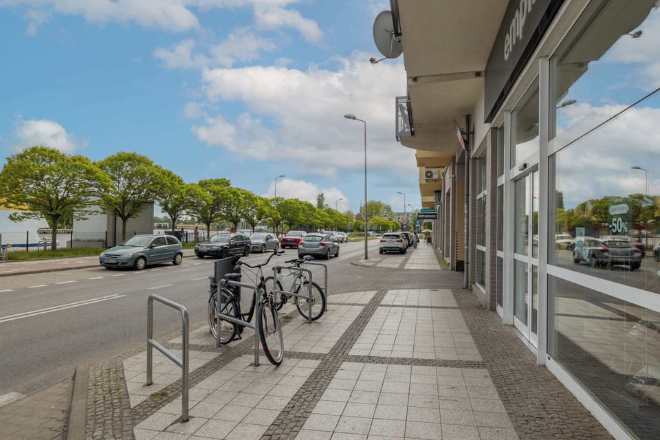 A view of the street with parked bikes, trees lining the path, and apartment buildings in the background.

