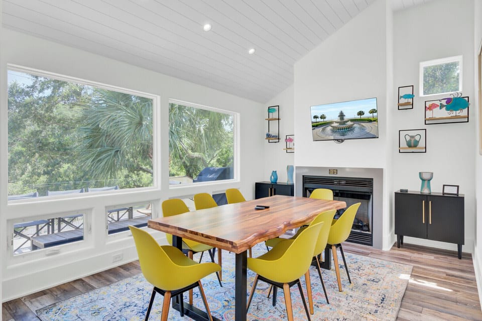 Your bright dining space features vibrant yellow chairs around a rustic wood table, with natural light streaming through windows overlooking tropical palms.