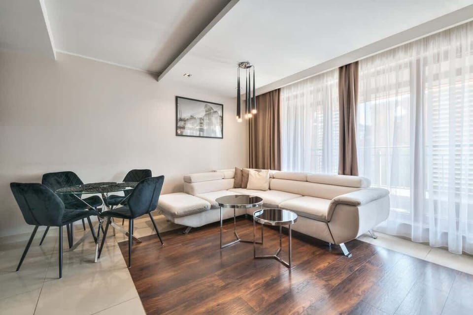 A spacious living room with a modern design, featuring a wooden floor, a glass coffee table, and comfortable seating. Large windows provide plenty of natural light.