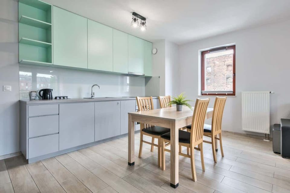A modern kitchen with sleek light gray cabinetry and a wooden dining table set with chairs. Large windows brighten the space.