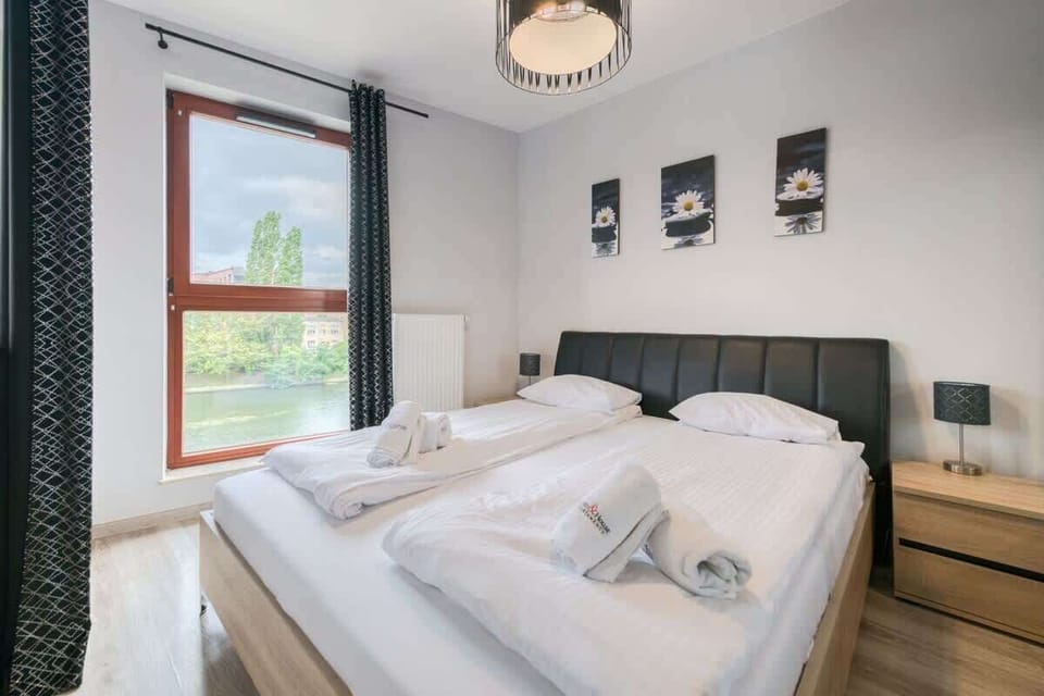 A cozy bedroom with a neatly made bed, white linens, and large windows overlooking greenery. The decor is modern yet welcoming.