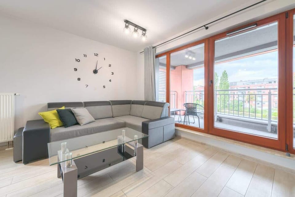 A bright, modern living room with a gray couch, a glass coffee table, and a dining area in the background. The room is well-lit and features contemporary decor.