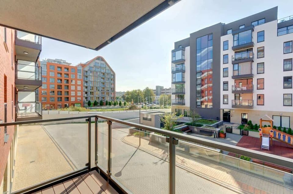The apartment's balcony overlooks modern buildings in an elegant urban neighborhood, with surrounding greenery and a children's playground. The architecture combines modernist forms with warm color accents, creating a pleasant and peaceful atmosphere