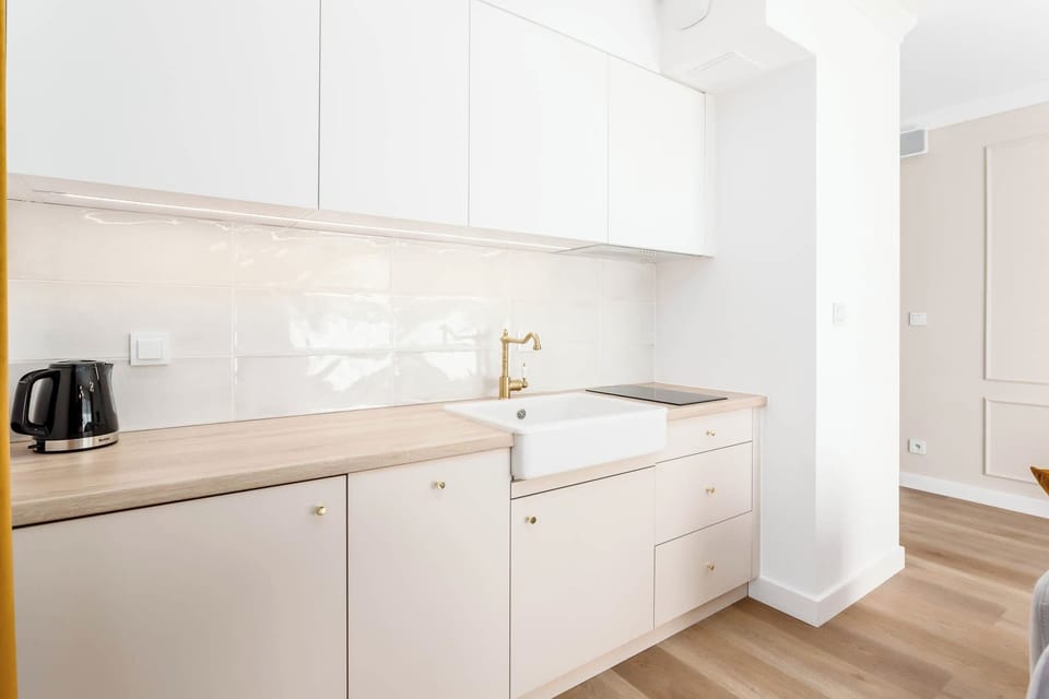 A bright, fully equipped kitchen with white cabinets and wooden flooring. It features essential appliances including a kettle and stovetop.