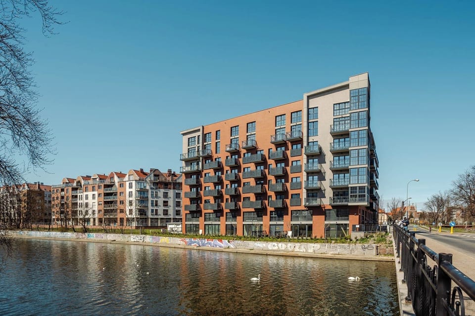 A scenic view of contemporary apartments along the water. The clear sky reflects beautifully on the river.
