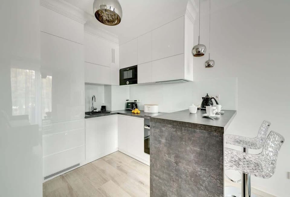 A sleek, modern kitchen with glossy white cabinetry, a minimalist design, a small black sink, built-in appliances, and a gray breakfast bar paired with transparent bar stools, complemented by metallic pendant lights.