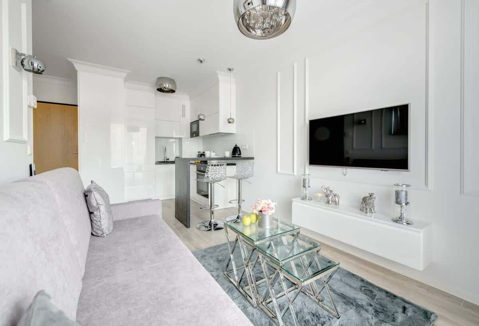 A modern open-plan living area with a sleek gray sofa, a glass coffee table, a minimalist kitchen with a breakfast bar, and a wall-mounted TV above a white console, all accented by elegant metallic decor and pendant lighting.