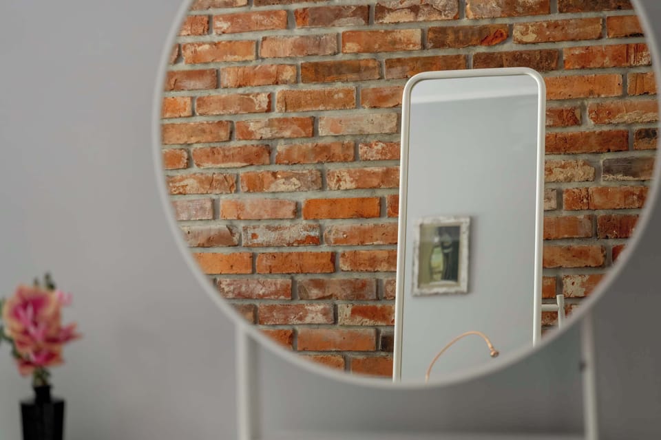 A round mirror mounted on a red brick wall above a vase of artificial flowers in a minimalist entryway.