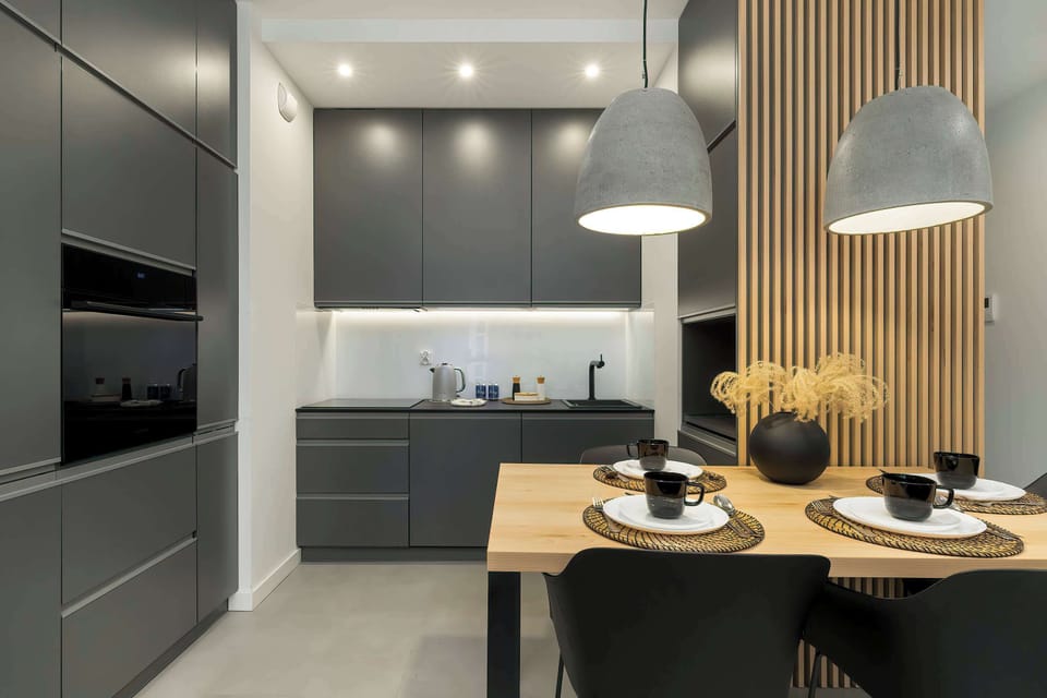 View of the kitchenette with dining area. Dark furniture contrasts with light walls and wooden panels. Coverings are set on the table, suggesting readiness for a meal.