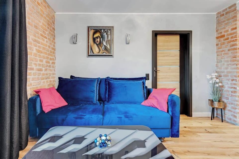 A fold-out sofa in a bold color, paired with decorative cushions, adds vibrancy to the space, while the brick wall serves as a striking backdrop for the minimalist furniture. 