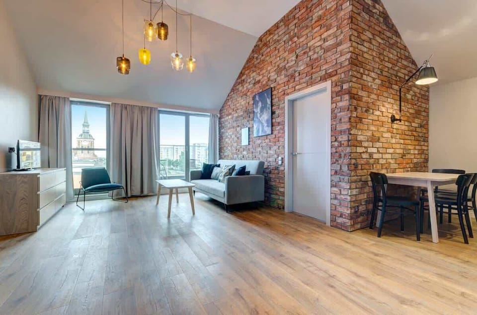 Large windows fill the room with natural light, showcasing industrial details like the brick wall. A designer coffee table and soft rug add warmth, making it the perfect place to unwind.