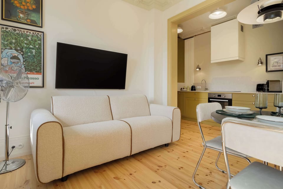 An open-plan living room with a small kitchen visible behind a cozy seating area and a wall-mounted TV.