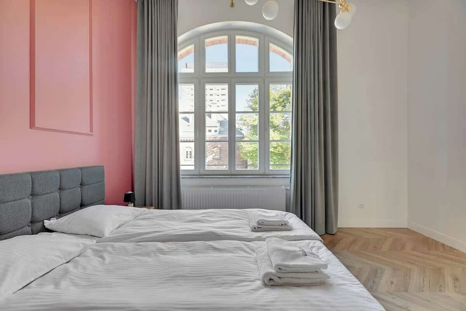Serene aesthetic, with a striking pink accent wall, plush bed linens, and soft gray curtains, all framed by abundant natural light through large windows, creating an inviting and relaxing atmosphere.
