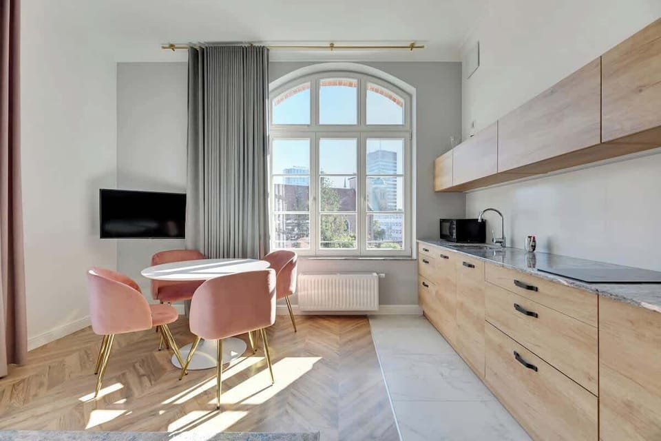 Refined, contemporary living space exudes elegance with its soft pastel accents, clean lines, and natural wood finishes, while large windows offer bright, airy views of the surrounding cityscape.