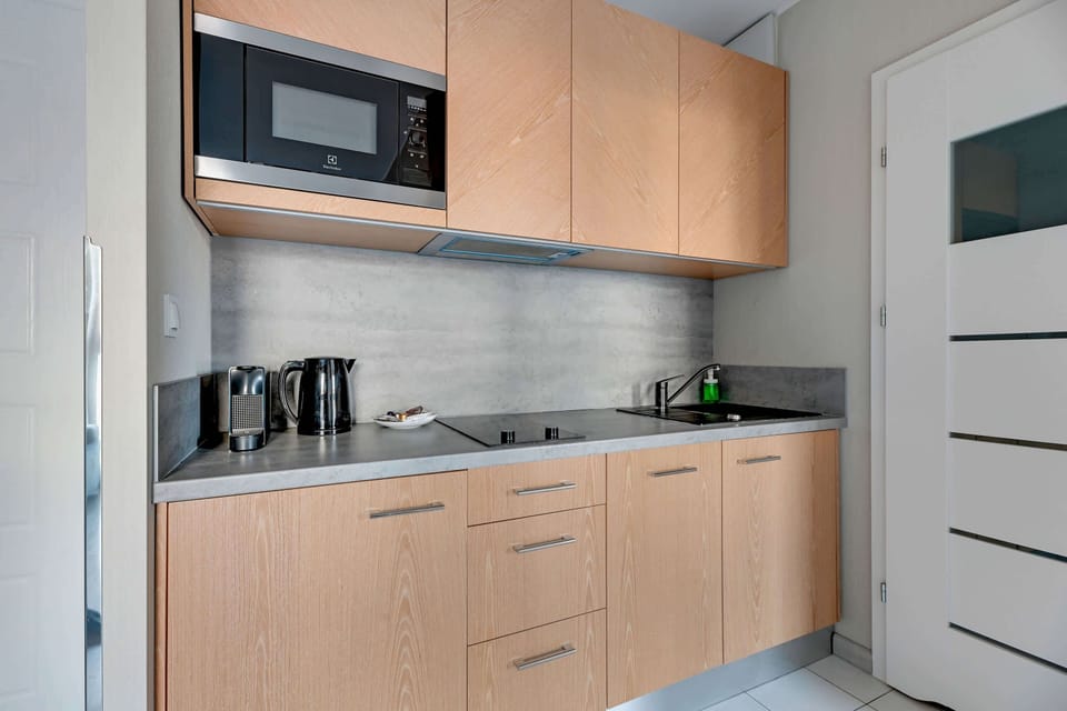 A compact kitchenette is fitted with wooden cabinets and a sleek gray backsplash. Appliances like a kettle and microwave are neatly arranged for convenience.