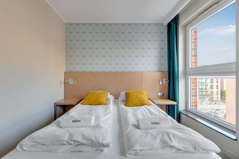 The bedroom offers a bright and cozy vibe with two beds adorned with white linens and colorful pillows. The large window provides a beautiful outdoor view.