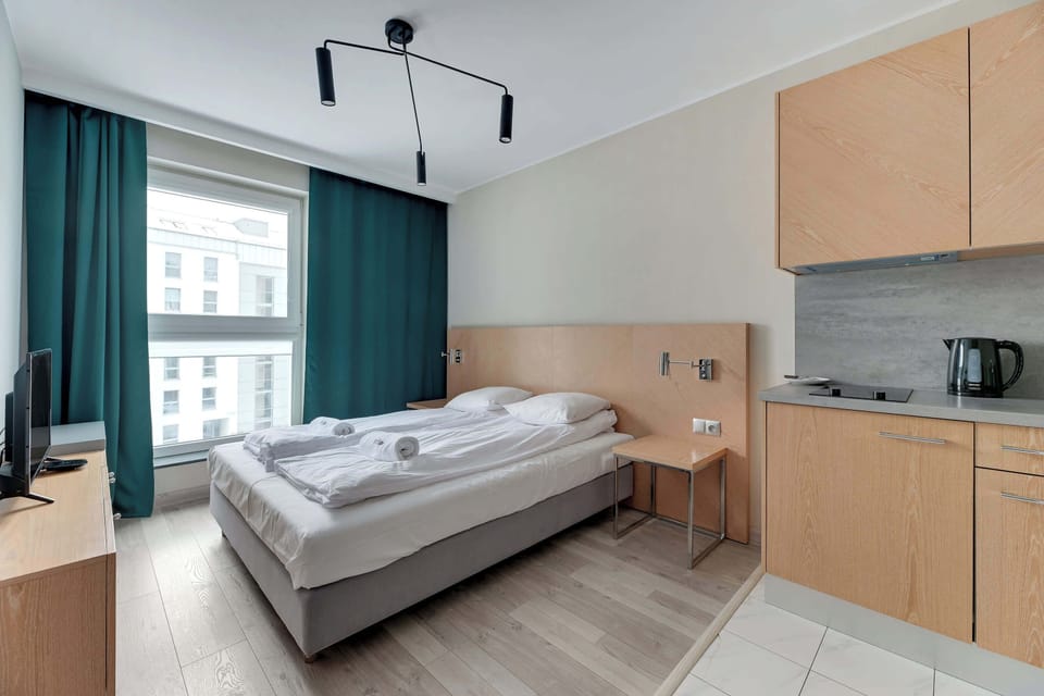 Studio, showcasing the bed with white linens and a wooden headboard. A large window framed with curtains allows ample light to fill the room.