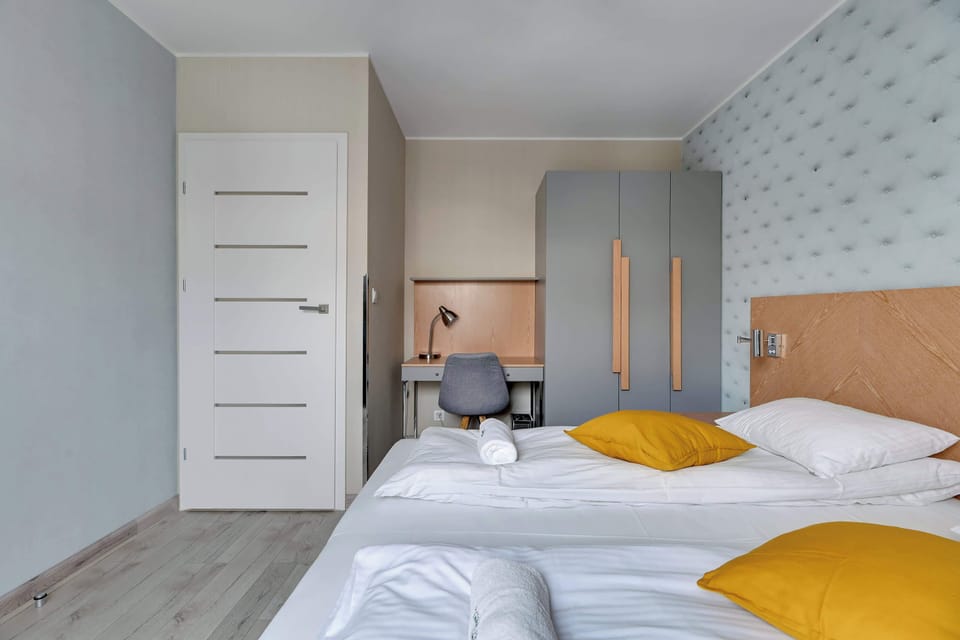The bedroom offers another view of the neatly made bed with yellow accent pillows and a spacious wardrobe. The patterned wallpaper adds character to the serene and well-lit room.