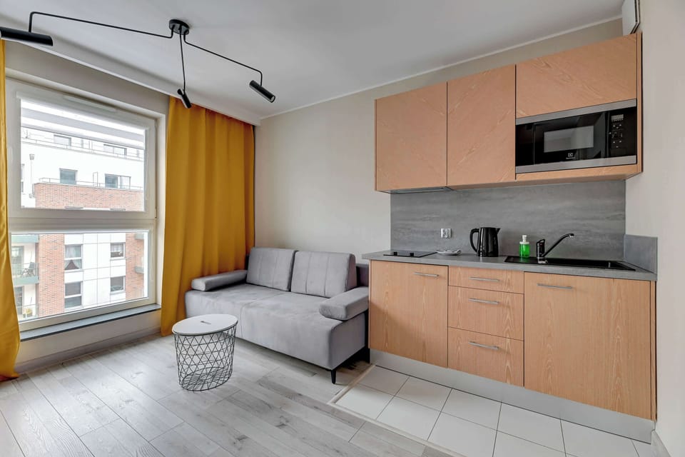 In the open-plan living area, a small kitchenette is visible alongside the sofa and a round white table. Yellow curtains and modern furnishings add warmth and practicality to the space.