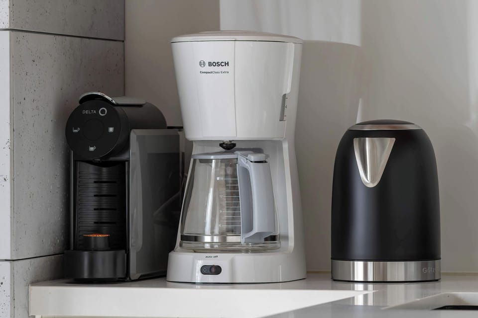 A kitchen appliance station with a coffee maker, kettle, and capsule espresso machine on a white counter.

