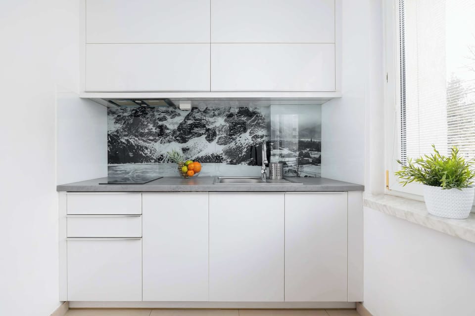 A kitchen with a minimalist design, featuring white cabinetry and a dark marble-patterned backsplash. A small potted plant adds a hint of greenery.