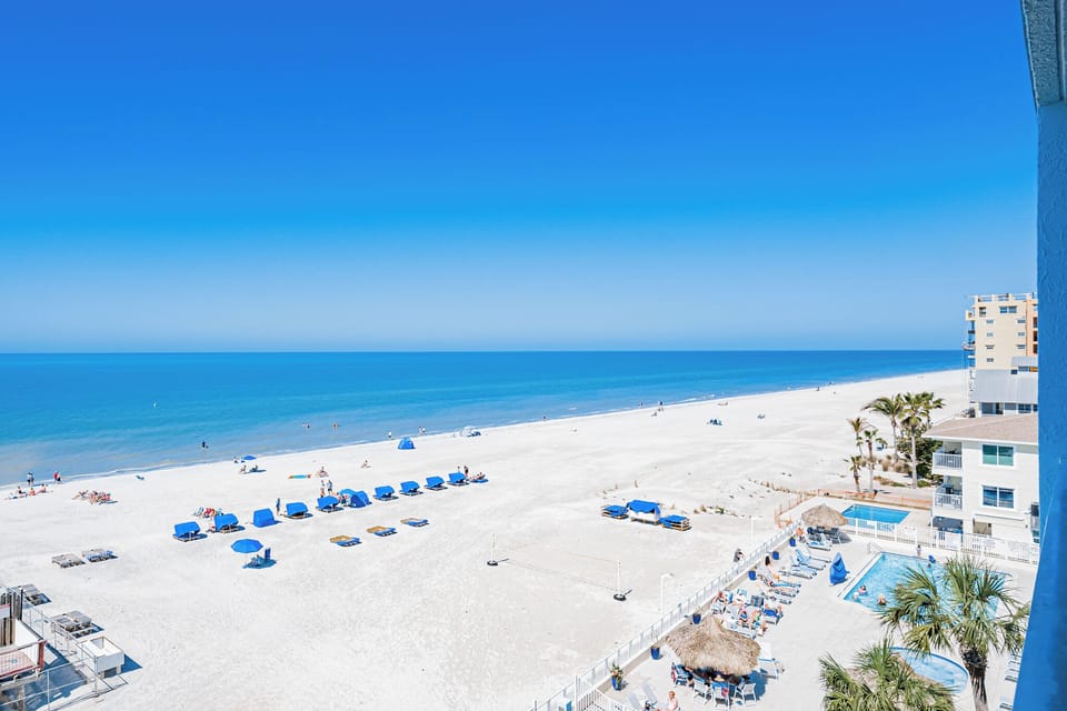 Pristine white sand beach stretches along crystal blue waters, with palm trees and beachfront buildings completing the coastal scene.