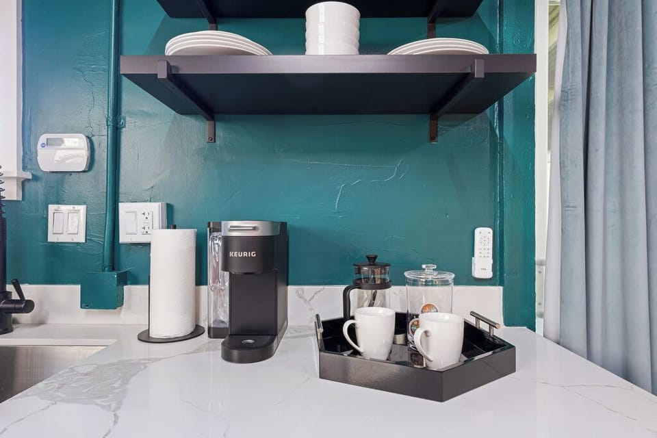 This cozy coffee nook features a sleek countertop with a Keurig machine, mugs, and condiments neatly arranged in a tray. Floating shelves above hold dishware, all set against a bold teal wall.