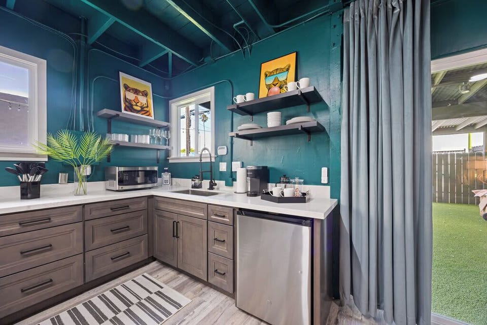 This cozy, vibrant kitchen showcases teal walls, modern open shelves, a mini fridge, and sleek gray cabinets. Bright lighting, playful artwork, and greenery add warmth and charm to the compact space.