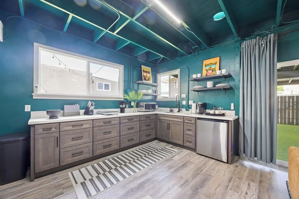 This modern kitchen features teal walls, sleek gray cabinetry, open shelving, and stainless steel appliances. Natural light fills the space, highlighting the clean design and stylish ceiling accents.